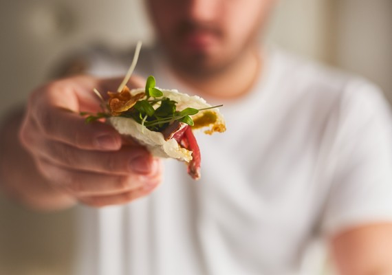 Homem a segurar, de braço esticado, no pão recheado com salada e outros ingredientes